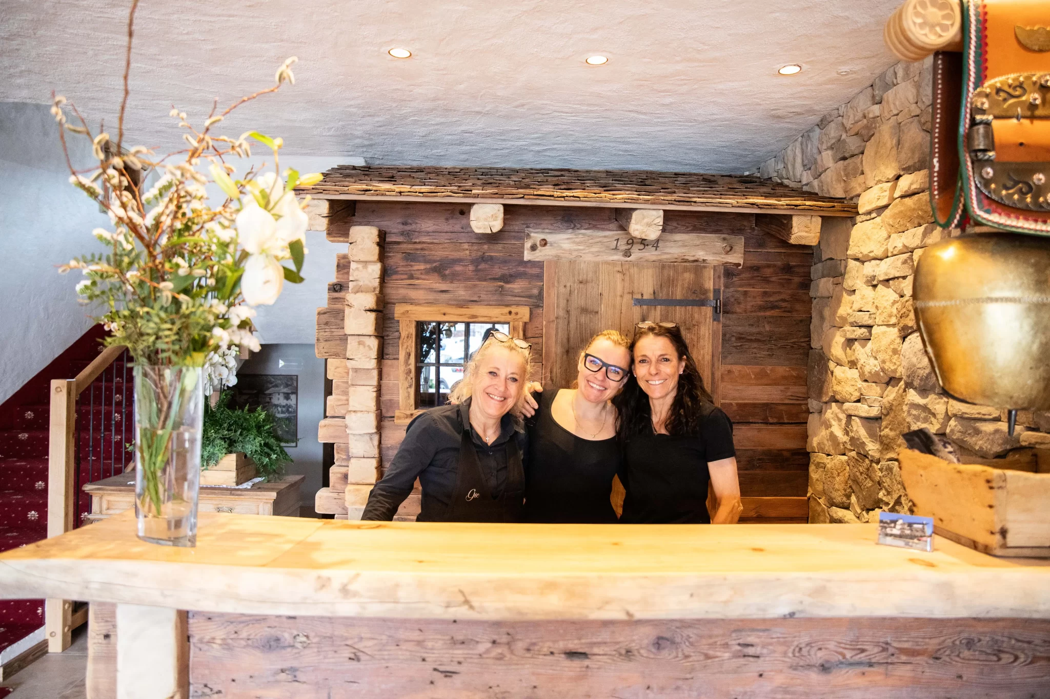 Tre persone sorridenti dietro un bancone di legno rustico con decorazioni floreali e pareti in pietra.