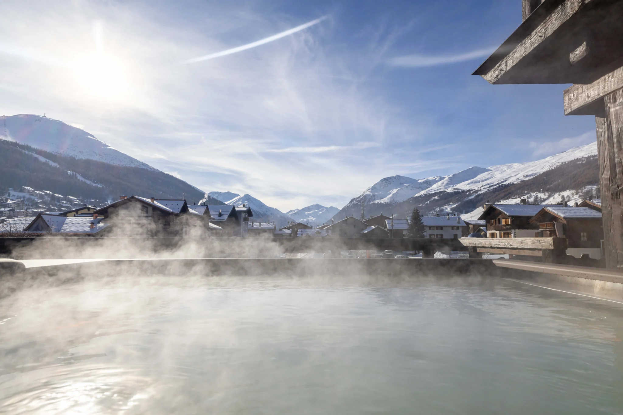 Vista di un paesaggio montano con vapore sopra una piscina calda, casette e montagne nevose sullo sfondo.