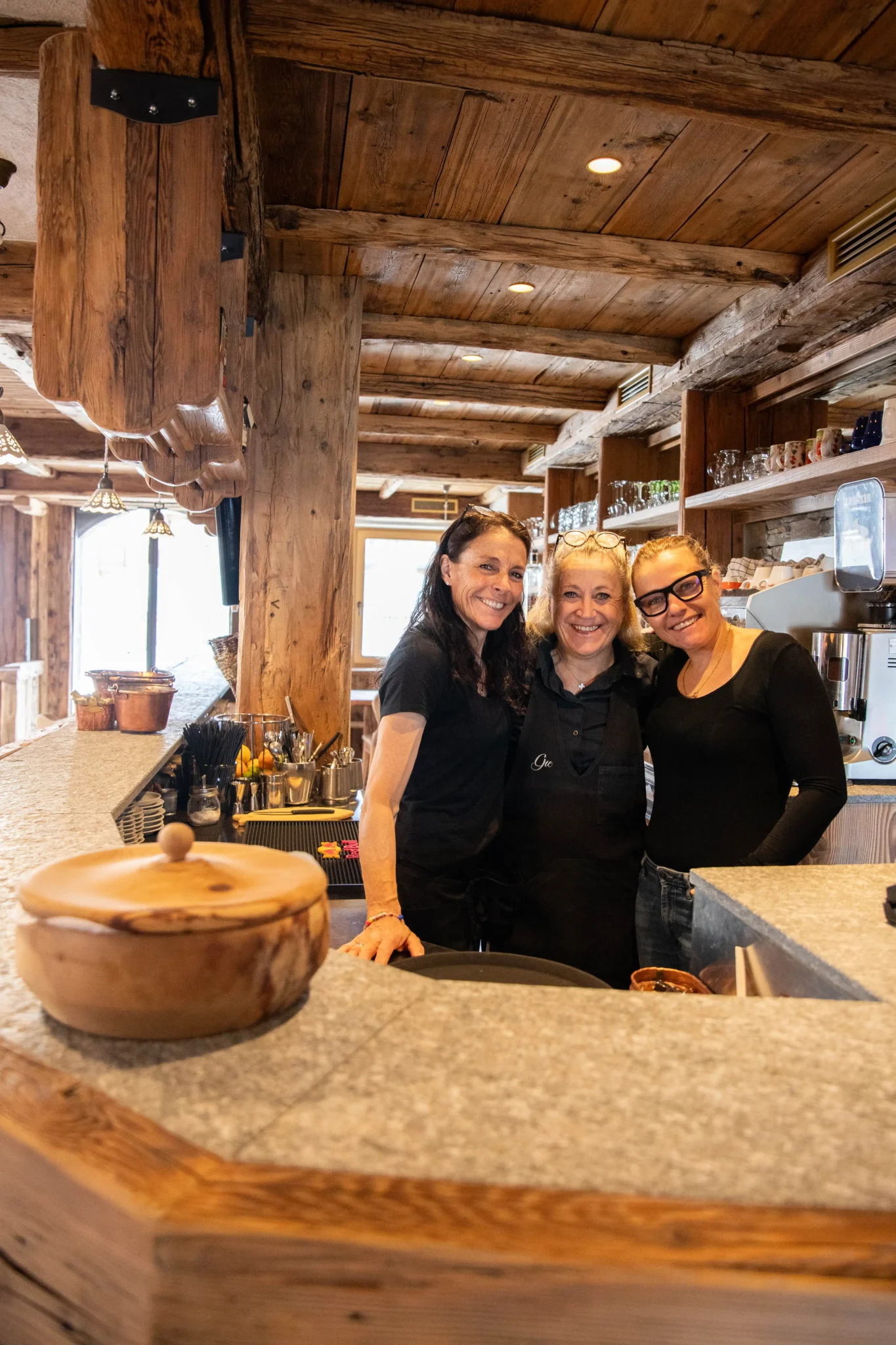 Tre donne sorridenti dietro un banco bar in un ambiente rustico in legno.