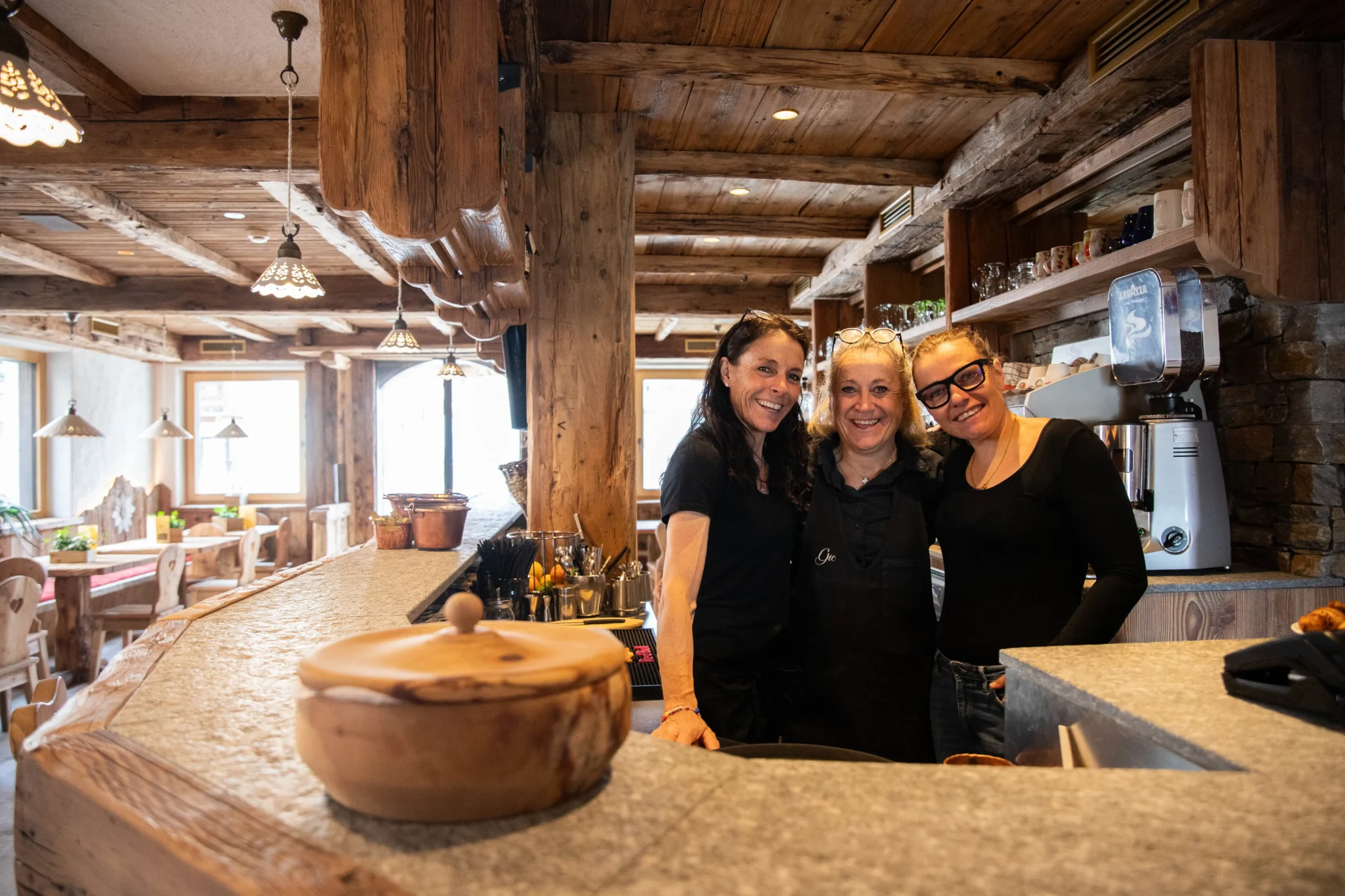 Tre donne sorridenti dietro il bancone di un accogliente bar in legno.