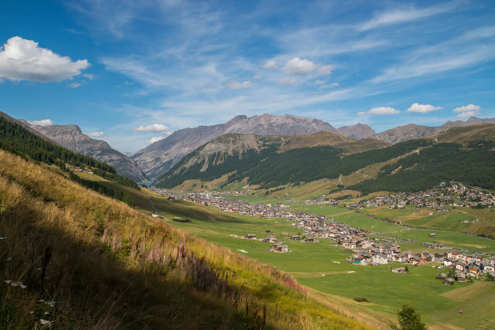 Vista panoramica di un villaggio alpino circondato da montagne e prati verdi sotto un cielo azzurro.