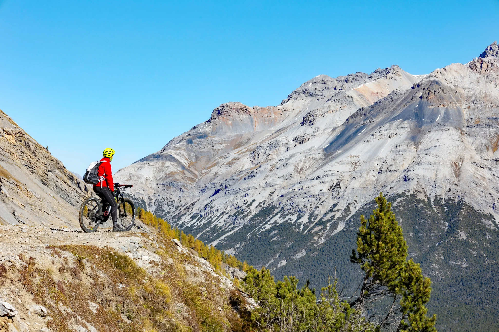 Ciclista con casco giallo e giacca rossa su un sentiero di montagna roccioso.