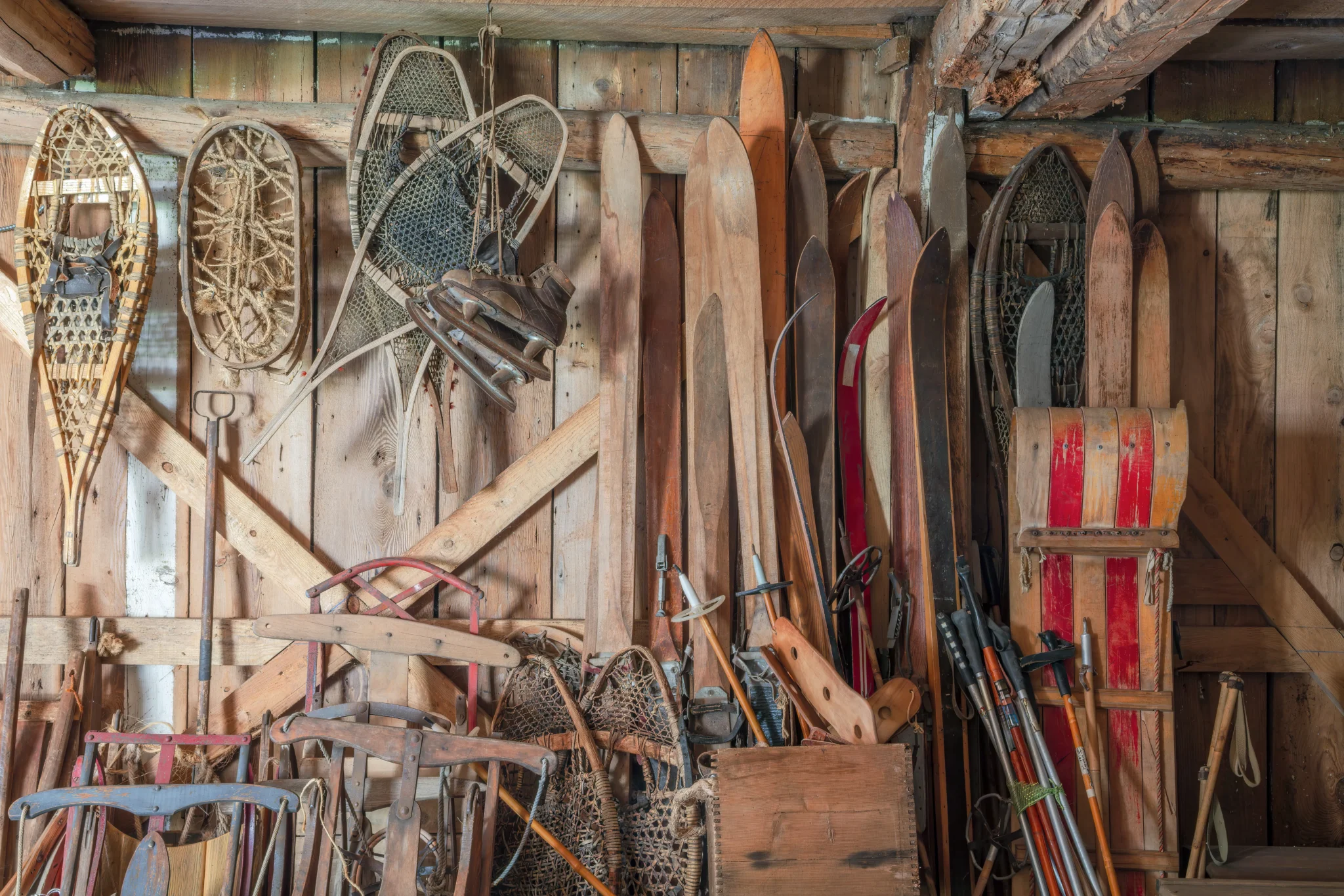Vecchia collezione di sci, racchette da neve e slittini in legno appesi su un muro di una baita rustica.