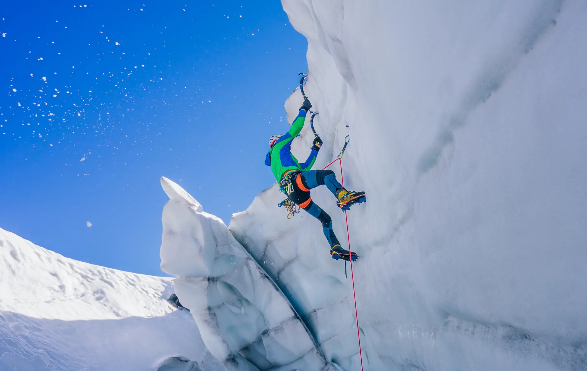 Alpinista impegnato nell'arrampicata su ghiaccio con piccozze e ramponi su una parete ghiacciata sotto un cielo azzurro.