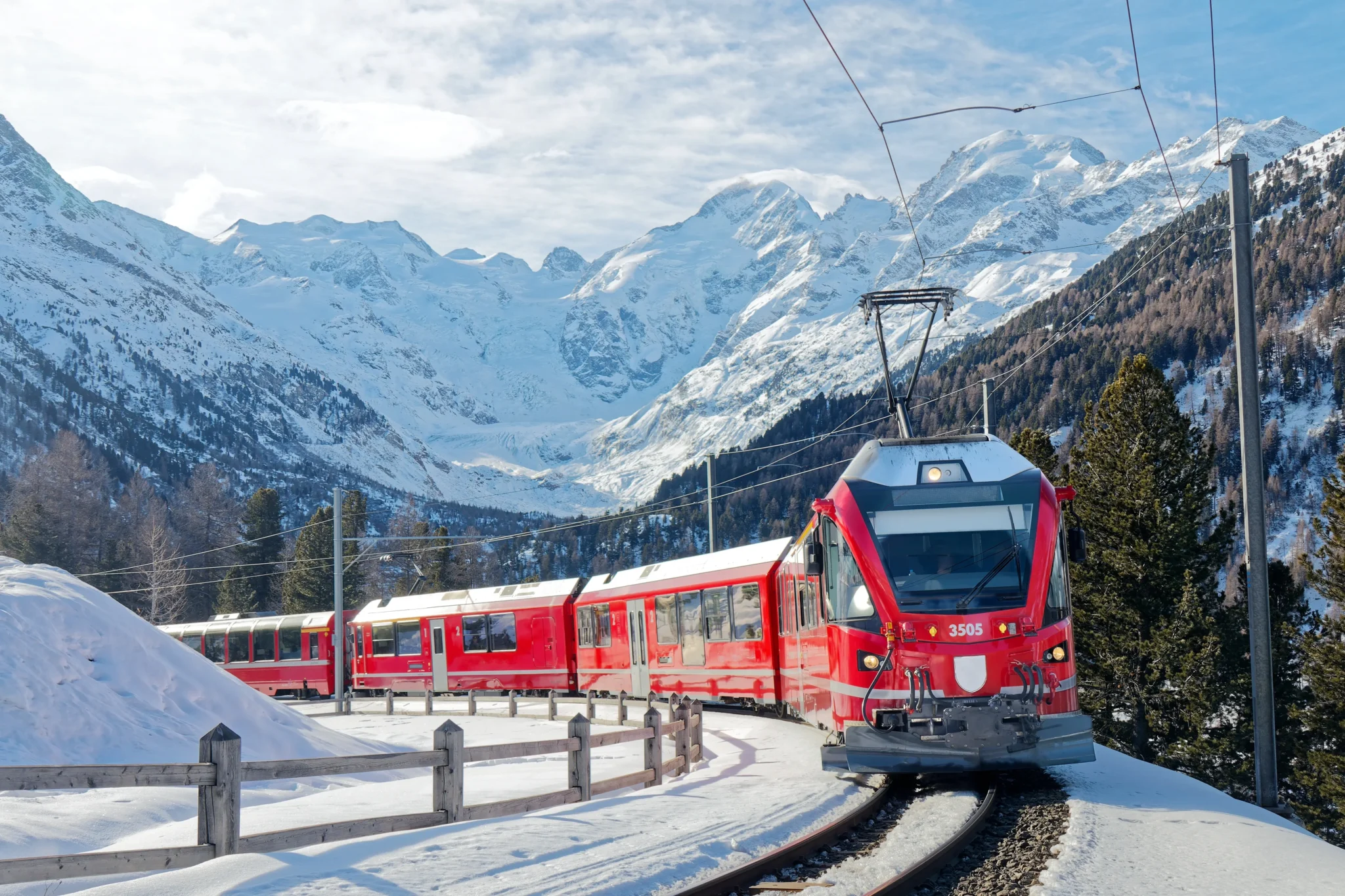 Treno rosso invernale attraversa un paesaggio alpino innevato con montagne sullo sfondo.